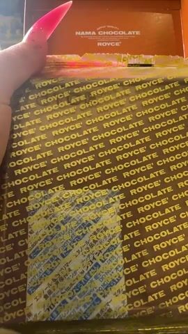 The video shows a close-up of a box of Royce' Nama Chocolate, a brand of Japanese chocolate. The box is predominantly brown with the word "ROYCE' CHOCOLATE" repeated in gold lettering. A Japanese language label is visible on the front of the box, likely containing product information or ingredients. The box is being opened by hands with long, bright pink artificial nails. The background suggests an indoor setting, though it is out of focus. The lighting appears to be artificial, possibly from indoor lights. The overall impression is one of anticipation and the unwrapping of a treat. The text visible on the box is "ROYCE' QUALITY" and "NAMA CHOCOLATE". The Japanese text is too small and blurry to be deciphered. There are no discernible time of day, weather, or specific location cues beyond the context of the product itself being from Japan. The emotions conveyed are primarily focused on the act of opening the chocolate.