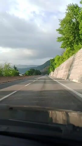 The video shows a first-person perspective of driving on a highway. The road has multiple lanes with white markings. On the right side of the road, there is a steep embankment with a concrete retaining wall and lush green trees. On the left side of the road, there are trees and some distant mountains under a cloudy sky. The view is forward-looking, showing the road ahead disappearing into the distance.