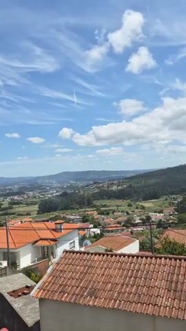 The video shows a daytime view of a neighborhood with homes featuring red tile roofs. Beyond the immediate houses, there is a landscape with green vegetation and distant hills. The sky is bright blue with scattered clouds. Loud noises resembling firecrackers or explosions are heard throughout the recording.