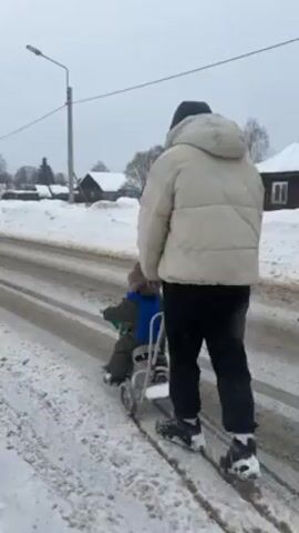 A man is pulling a child on a small wheeled snow sled. The man wears a light-colored puffer jacket, black pants, and black sneakers. The child is bundled up in a green snowsuit and a blue scarf, seated on the sled, which has a metal frame and small wheels. They are walking on a snow-covered road with tracks from vehicles. The environment shows a rural setting with houses visible in the background and snow-covered yards. A street light pole is visible on the side of the road. The scene is overcast and the ground is a mix of snow and slush.