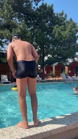 A person in black swim trunks jumps into a swimming pool from the edge. A yellow inflatable ring is visible in the water. There are lounge chairs and trees in the background. Another person is in the pool to the right.