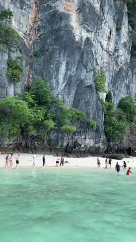 The video opens with a view of a rocky cliff face covered in lush green foliage. Below the cliff is a white sandy beach where several people are scattered. Some are standing on the beach, while others are wading in the clear, turquoise water. In the foreground, the water is calm and inviting. As the video progresses, a white boat with blue lettering appears on the right side of the screen. People are seen getting on and off the boat, suggesting it is a mode of transportation to or from the beach. The overall scene depicts a tropical beach location with people enjoying recreational activities by the water and amidst natural beauty.