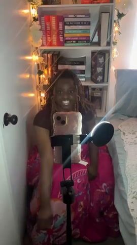 The video shows a woman sitting in a small space, facing the camera. She has curly hair in braids, wears a brown t-shirt, and is covered with a pink patterned cloth. She is smiling at the camera. In front of her is a smartphone mounted on a stand, with a microphone attached. To her left, there is a white wall decorated with fairy lights and artificial flowers. To her right, there is a bed covered with a light blue blanket. Behind her, there is a white bookcase filled with books. The room is well-lit, and the woman appears to be recording a video or engaging in a video call.