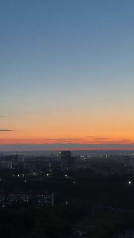 The video presents an aerial view of a city at dusk, transitioning into night. The sky displays a striking gradient, moving from a deep blue at the zenith to vibrant orange and red hues along the horizon, indicative of a sunset. A solitary bright star or planet is visible in the upper left quadrant of the sky initially.

The camera, positioned at a high vantage point, slowly pans downwards and rotates slightly, revealing the urban landscape below. As it descends, the city lights become more prominent, illuminating streets, buildings, and other structures. The scene encompasses a mix of multi-story residential buildings, areas with dense tree cover, and commercial establishments. Roads are visible, with both stationary and moving vehicles. Towards the end of the clip, a gas station with distinct red and white signage comes into clearer view, along with a parking lot occupied by numerous cars. The overall impression is a serene and expansive cityscape as evening settles over the area.