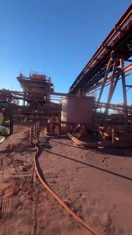 The video shows an industrial site under a clear blue sky. The ground is covered in reddish-brown dirt, with some sandy patches and footprints visible. Several large, rust-colored metal structures dominate the scene, including conveyor belts supported by a network of beams and columns. In the mid-ground, a cylindrical tank with some machinery attached sits on the ground. Further back, there is a multi-level processing plant or building with more conveyor belts. The lighting suggests it is either early morning or late afternoon, casting long shadows. The overall impression is of a mining or ore processing facility. No people are visible in the footage.