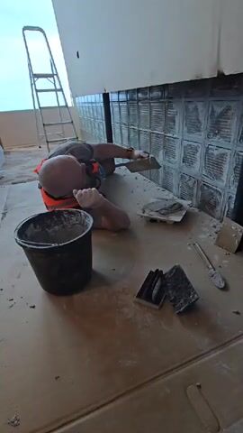 A man in an orange safety vest, gray gloves, and a watch on his left wrist is shown working on a wall constructed of glass blocks. He is lying on his side on a surface covered with brown paper, using a yellow and black trowel to apply a gray substance to the glass blocks. A black bucket containing a similar gray material sits beside him. To his right, on a square white tile, is a pile of the gray material. Near the tile, there are two black objects and a trowel partially coated in the gray material. Behind him, a ladder is visible in the corner of the room, and a large window allows natural light to illuminate the space. The overall impression is that of a construction or renovation scene.