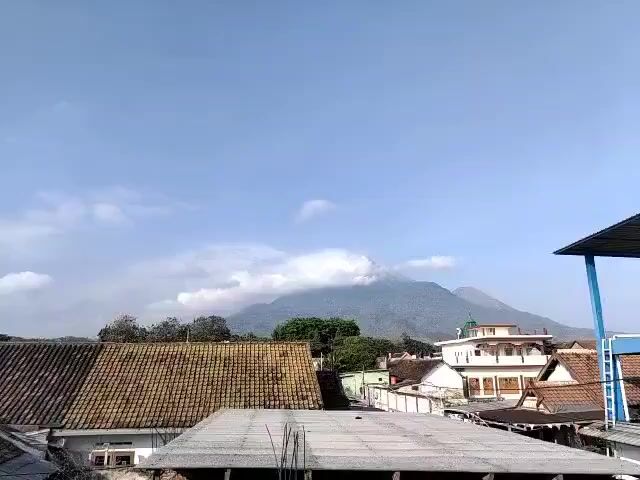 The video shows a panoramic view of a village with a mountain in the background. The sky is clear with a few white clouds. In the foreground, there are several houses with tiled roofs. To the right, there is a blue water tank on a platform. The mountain is covered in mist at the top. The video is shot from a high vantage point, possibly a rooftop. There are no people visible in the video. The overall scene is peaceful and scenic.