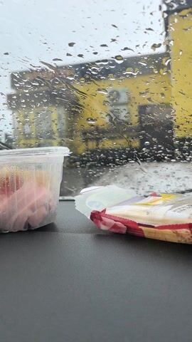 A static shot from inside a car shows a plastic container of sliced ham and a partially opened package of crackers on the dashboard. The windshield is covered in raindrops, obscuring a yellow building in the background. The light is overcast, suggesting a cloudy or rainy day. A song is playing.