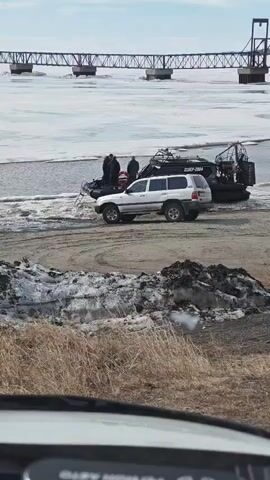 The scene shows a partially frozen river landscape with a bridge in the background. On the riverbank, there is a white SUV parked next to two black airboats. Several people are standing near the boats, seemingly engaged in some activity. The foreground includes patches of snow and dry grass. The day appears overcast.