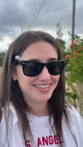 A woman with medium-length brown hair, wearing Ray-Ban sunglasses, a white button-down shirt, and a white t-shirt with a partial image of the word "ANGELES" is shown in a close-up shot.  She is outdoors with a blurred background of greenery, an overcast sky, and what appears to be a utility pole. She smiles, then blows on a small object held in her hand. The object appears to be a dandelion seed head on a stick. She looks directly at the camera while blowing on it. She has a small flower tucked behind her ear, and her nails are neatly manicured with a light pink color.