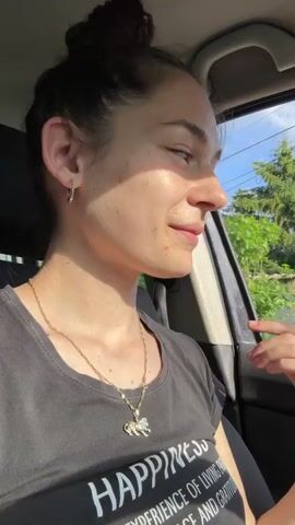 A young woman is sitting in the driver’s seat of a car. She’s wearing a black t-shirt with the word "HAPPINESS" printed on it, along with a longer phrase. She has a gold necklace with a pendant. Her hair is pulled back in a bun and she’s wearing hoop earrings. She smiles at the camera, then turns her head to look out the side window. She raises her hand and waves. The car is parked, with green trees and buildings visible through the window. It's a sunny day.