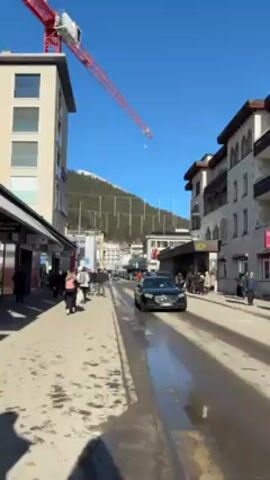 The video shows a street scene in a town, likely a resort town. The street is wet, with patches of melted snow or ice. There are pedestrians walking on the sidewalks on either side of the street. A black car drives down the street. Buildings line the street, and in the background, there is a mountain covered with trees, some with snow at the very top. A construction crane is visible above one of the buildings. The sky is clear and blue. Flags are visible in front of a building.