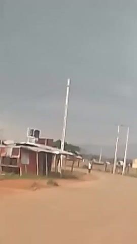 A person is walking on a dirt road. There are some small buildings and utility poles on the side of the road. The sky is overcast.
