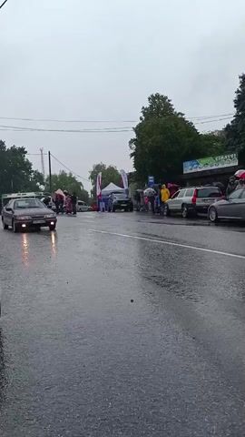 The video shows a street scene in rainy weather, with several cars and a few people visible. A dark-colored sedan is driving down the wet road in the foreground, its headlights on. In the background, there are more cars parked along the side of the road, and some people are standing under umbrellas. There are also some tents and booths set up, suggesting an outdoor event. The sky is overcast and gray.