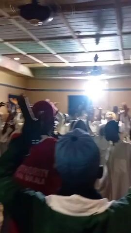 A group of people are gathered in a room with a patterned ceiling and beige walls. Many of the people are wearing white garments and head coverings, suggesting a religious gathering. Some individuals are raising their hands in the air, possibly in prayer or praise. One person in the foreground wears a green jacket and a blue and white patterned hat. Another person in the center wears a red shirt with white lettering visible on the back. A bright light illuminates the scene from the right side, casting some shadows and making it difficult to see details in that area. The overall atmosphere appears to be one of communal participation and spiritual engagement.