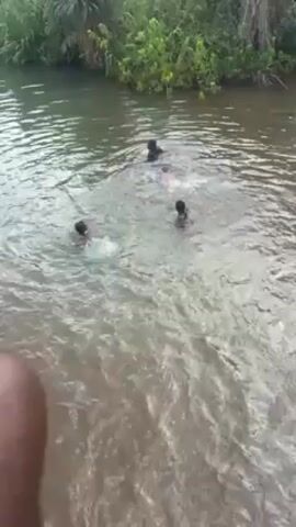 Four people are swimming in a river or stream. The water is murky, and lush green vegetation lines the banks. The individuals are clustered together, splashing around in the water. One of the swimmers is wearing a green top. A person holding the camera is filming the scene from what appears to be a bridge or an elevated position.