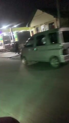 The video shows a nighttime view of a gas station. The gas station has bright, colorful lights illuminating the pumps and the "BEST PARK OIL & GAS" sign. Several cars are visible at the gas station, some appearing to be fueling. There are also a few people walking around the gas station. The video is shaky, suggesting it was filmed from a moving vehicle. The road in front of the gas station is visible and appears to be paved. The overall impression is a busy, well-lit gas station at night.