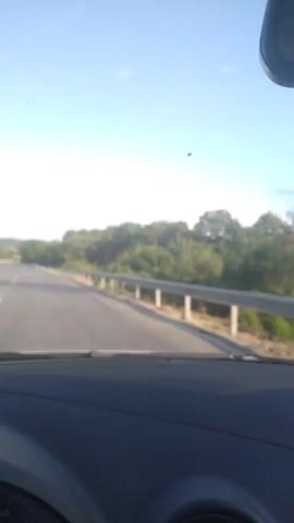 The video shows a point-of-view shot from inside a car driving on a highway. The road is asphalt with white lane markings. Guardrails are visible on the right side of the road, and trees line the sides of the highway. The sky is blue with some scattered clouds. In the second clip, a hand with pink nail polish is visible on the steering wheel. The car appears to be traveling at a moderate speed.