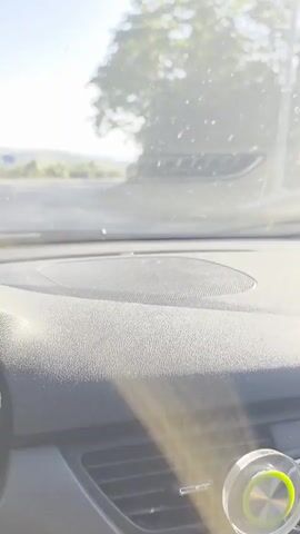 The video is filmed from inside a car, looking out the windshield. The car is driving on a paved road that curves to the right. On the left side of the road, there is a field with some vegetation and a signpost with blue arrows. On the right side of the road, there is a wooded area with trees. The sky is clear and blue. Two other cars are visible on the road, one in the distance on the right and another approaching on the left. The dashboard of the car, including a speaker and air vents, is visible in the foreground.