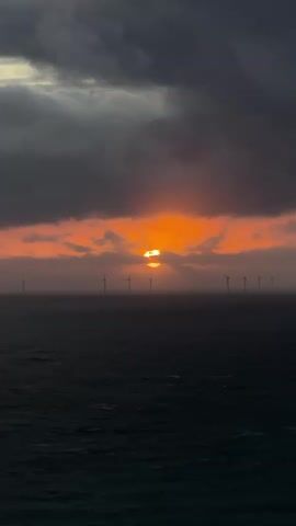 The video captures a dramatic seascape at what appears to be either sunrise or sunset. The dominant feature is a dark, churning ocean under a heavily clouded sky. The clouds are a mix of dark gray and ominous tones, with a break in the middle revealing a brilliant orange and yellow sky where the sun is positioned. The sun, partially obscured by clouds, casts a bright, intense glow, reflecting a shimmering path on the water directly below it.

Stretching across the horizon, silhouetted against the colorful sky, is a line of offshore wind turbines. Their dark forms stand in stark contrast to the glowing sun behind them, appearing as small, uniform structures in the vastness of the sea. The camera remains static, offering a wide shot of the scene. The overall atmosphere is one of natural power and beauty, with the dark, stormy clouds contrasting sharply with the fiery glow of the sun. The sound of wind is distinctly audible throughout the video, reinforcing the sense of a blustery day at sea. No people or text are visible.