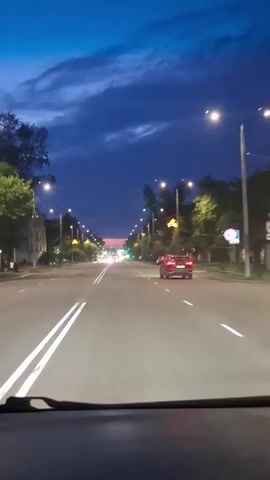 A car drives down a wide asphalt road at dusk. The sky is a deep blue with dark clouds. Streetlights line both sides of the road, illuminating the trees and buildings. A red car is visible ahead in the right lane. The video is shot from inside another vehicle, showing the dashboard and windshield wipers at the bottom of the frame. Traffic lights in the distance indicate an intersection.