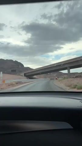 This video captures a drive through a highway overpass in Boulder City, United States. The camera is positioned inside a vehicle, looking out through the windshield. The scene is set in a desert-like environment with arid, rocky hills on either side of the road. The sky is overcast with dramatic clouds, suggesting a possibly stormy or cloudy day, but the overall lighting is bright, indicating it's daytime.

The primary activity is driving on a multi-lane highway that curves gently. An elevated highway overpass dominates the foreground, casting a shadow over the road below. Several cars are visible on the highway, moving in the same direction as the camera vehicle. Traffic lights are also visible, with some showing red. The surrounding landscape is characterized by dry, reddish-brown earth and sparse vegetation. There are also guardrails, road signs, and concrete barriers typical of a highway infrastructure.

Notable details include the concrete structure of the overpass and the vast, open desert scenery. The weather appears to be cloudy, contributing to a somewhat dramatic and subdued atmosphere, though no strong emotions are conveyed by the imagery itself. The location is identified as Boulder City, United States. No people are distinctly visible, as the focus is on the driving experience and the environment. There are no discernible words on any signs that are clear enough to read.