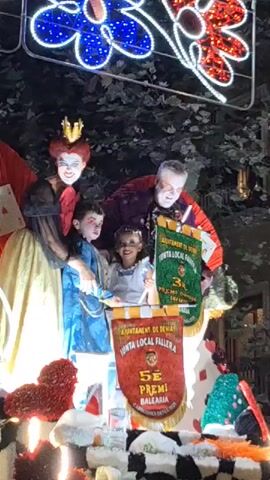 The video shows a close-up view of a parade float with people dressed in costumes. In the center, a woman in a Queen of Hearts costume, complete with a crown and dramatic makeup, is posing and interacting with the camera. To her left, a man dressed as a King of Hearts also has theatrical makeup and appears to be looking at the camera. Between them, two children are dressed as Alice in Wonderland characters. The float itself is decorated with large playing cards, red heart shapes made of what appears to be floral material, and a black and white checkered pattern. Banners are visible on the float, one with text in Spanish, indicating it is a winner of some kind of award or competition from the "Junta Local Fallera". The lighting suggests it is nighttime or indoors, with bright lights illuminating the float and costumes.