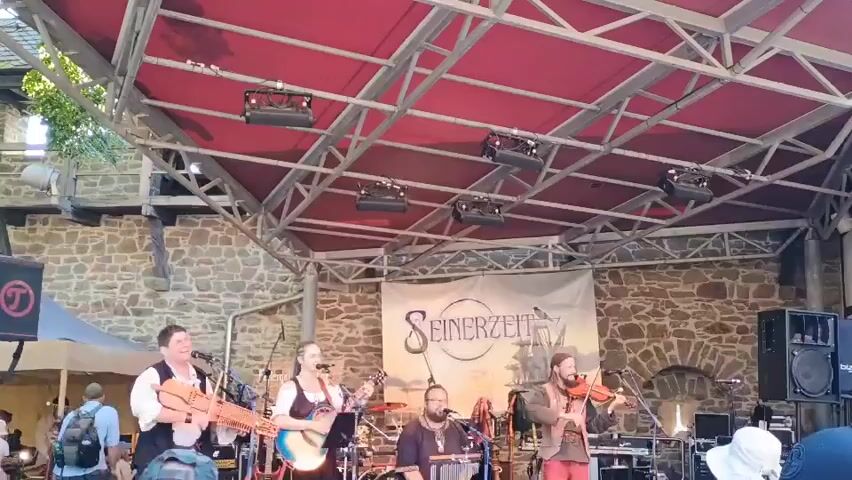 A band performs on a stage outdoors in front of a stone building. The stage has a red canopy and is supported by metal beams. Several lights are attached to the beams above the stage. In the background, a banner reads "Seinerzeit" with an image of a ship. There are four musicians on stage. On the left, a man plays a hurdy-gurdy. Next to him, a woman sings into a microphone and plays a guitar. In the center, a man plays a keyboard. On the right, a man plays the violin. Other instruments and equipment are visible on stage, including drums and amplifiers. Several people are in the audience, and tents are set up to the left of the stage. The overall scene appears to be a festival or outdoor concert.