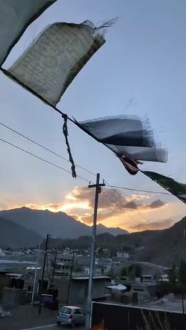 The video captures prayer flags fluttering in the wind against a backdrop of a dusky sky and mountainous terrain. The flags, a mix of white, black, and colorful fabrics, are strung across wires, their edges frayed and tattered by the wind. In the distance, mountains with soft, rounded peaks are silhouetted against a sky painted with hues of orange and yellow from the setting sun. Below the flags, a small town or village is visible, with buildings clustered together. A few utility poles and wires are also present in the scene. The overall impression is one of a serene, natural landscape touched by human presence.