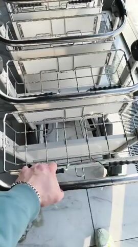 A person pulls a luggage cart away from a stack of other luggage carts in an airport. The cart is made of chrome with a wire basket and white shelving inside. The person has a bracelet and is wearing a light blue sweater. The floor is white and tiled with grey veining, and people and luggage can be seen in the background.