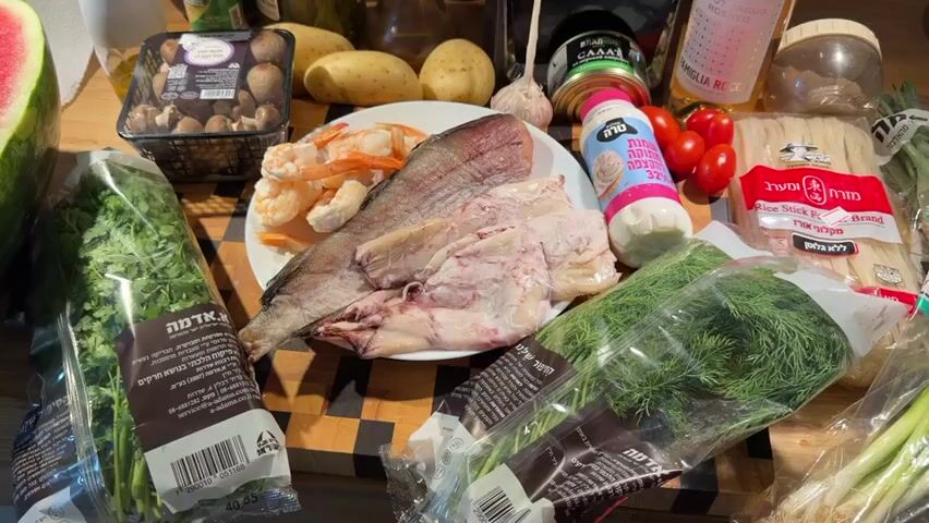 The video displays a close-up shot of various food ingredients arranged on a wooden surface. The scene is well-lit, suggesting it's likely indoors, possibly a kitchen. In the foreground, there are packages of fresh herbs, including what appears to be cilantro and dill. A plate in the center holds a raw fish fillet, several shrimp, and some other unidentifiable pieces of seafood. To the left of the plate is a slice of watermelon. Potatoes and a head of garlic are visible behind the plate, along with a container of mushrooms. A can of what seems to be salad is also present, next to a bottle of pink wine and a small bottle of olive oil. A small container of cherry tomatoes and a bottle of what looks like a creamy sauce are also in the frame. Towards the right, there's a package of rice stick noodles and some green onions. A blender is visible in the background, to the left of the wine bottle. The overall impression is that of ingredients being prepared for a meal.
