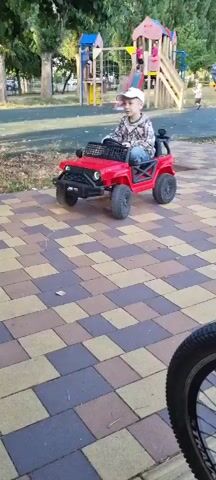 A young boy wearing a plaid shirt, blue jeans, and a white baseball cap is sitting in a red toy Jeep. The boy is in a park-like setting with trees and a playground in the background. The ground is paved with colorful interlocking tiles. The boy is holding the steering wheel and looking around. He briefly points to his left. The Jeep has a black grille and black wheels. A portion of a bicycle wheel is visible in the foreground on the right side of the frame.