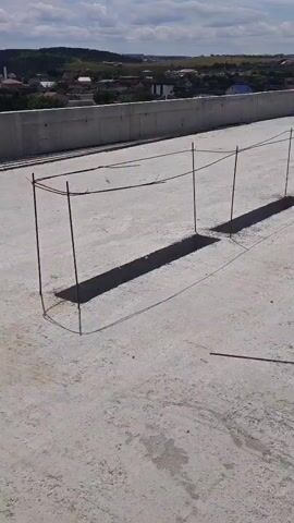 The video shows a rooftop construction site under a bright sky with scattered clouds. In the foreground, a rough concrete surface is visible, with several rectangular cutouts and rebar structures protruding from the ground. These rebar structures appear to be forming the outlines of future concrete elements. In the background, a low concrete wall runs along the edge of the rooftop, and beyond that, a landscape of hills and buildings can be seen under the sunlight. The overall impression is a construction project in progress, with the concrete slab of the rooftop laid and reinforcement work underway.