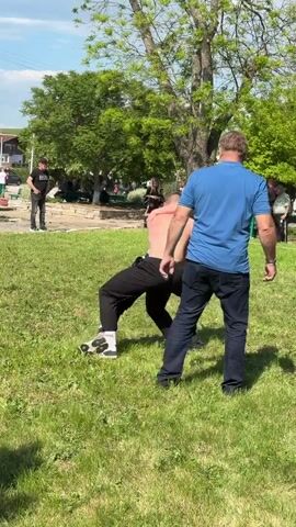 In a grassy outdoor area, two shirtless men are wrestling. They are on the ground, grappling with each other. One man is wearing black athletic pants with a white stripe, while the other is wearing black pants. A man in a blue shirt and dark jeans stands nearby, watching the wrestling. In the background, other people are sitting and standing around, observing the scene. There are trees and a building visible in the distance. The weather appears to be sunny.