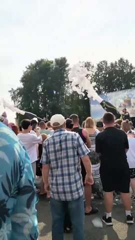 A group of people are gathered outdoors, likely at an event or festival. In the foreground, the backs of several individuals are visible, including a man in a plaid shirt and a woman in a white top with a black and white patterned skirt. The crowd is facing a stage area where a man is operating a foam machine, spraying large amounts of white foam into the air. Some people in the crowd are reaching up, seemingly enjoying the foam. In the background, trees and a backdrop with a painted image of buildings, possibly a church, are visible. The sky is bright and appears to be daytime.