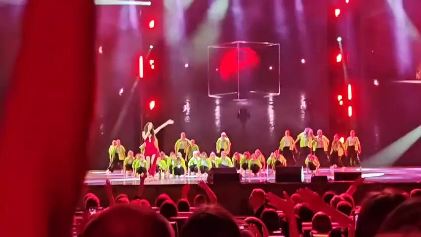 A stage performance is captured, bathed in red and white stage lights. A central female performer in a red dress and a male performer in a black cap and outfit are on the stage, interacting. Behind them, a large group of performers, mostly young people, are dressed in bright green jackets and black pants, positioned in rows. They are in various poses, some crouching and some standing. A large, translucent cube structure with a red glowing sphere inside is visible in the background, suspended above the stage. The audience is visible in the foreground, with their hands raised, suggesting they are engaged and enthusiastic.