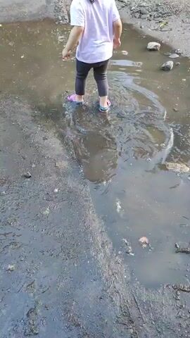 A child, seen from behind, is standing in a puddle of murky water. The child is wearing a light-colored shirt, dark pants, and pink and blue shoes. The ground around the puddle is wet and muddy. The water is disturbed, suggesting the child is moving or playing in it.