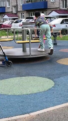 The video shows a playground scene. In the foreground, a blue and black scooter is parked. The ground is covered with a colorful rubberized surface featuring blue, green, and yellow circles. A carousel with yellow seats and a gray metal structure is central to the scene. A boy wearing a black cap, a t-shirt with a graphic print, and light green pants is standing on the carousel, facing away from the camera and appearing to push it. To the right of the carousel, a young girl wearing a pink cap, a tie-dye shirt, and pink leggings is holding a baby dressed in blue. The girl is facing the carousel and the baby is looking towards the carousel. Another child is partially visible on the right edge of the frame, also facing the carousel. In the background, there is a multi-story building and parked cars. The overall impression is a snapshot of children playing on a playground.