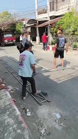 Several people are gathered on a street in front of houses, possibly setting up or dismantling equipment. One person in a light blue t-shirt and black pants is stepping on a metal frame on the road. Another person in a black t-shirt and black shorts is also holding a metal pole. In the background, other individuals are visible, some near a red truck, and others near a building with a balcony. The scene appears to be outdoors on a sunny day, with debris scattered on the road and sidewalk. Some metal scaffolding or frames are laid out on the street, and there are also power lines visible overhead.
