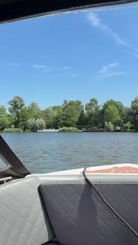 A video shot from the interior of a boat looking out at a body of water. The sky is blue with some white clouds. Lush green trees line the shore of the water. Several boats are visible docked along the shore. The interior of the boat shows gray quilted seats and a rope coiled on the floor. A clear plastic covering is partially visible on the right side of the frame. The boat appears to be moving slowly through the water.