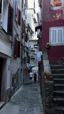 The video shows a narrow cobblestone alleyway between buildings in what appears to be a Mediterranean town. The buildings are made of stone and plaster, with red shutters and windows. Laundry is hanging from lines stretched across the alley. On the right side of the alley, there is a stone staircase leading upwards. An orange cat is sitting on the wall next to the staircase, looking at the camera. Two people, one wearing a white shirt and the other a dark dress, are walking away from the camera down the alley. The lighting suggests it is daytime, possibly late afternoon.