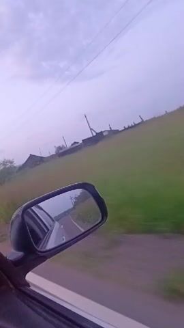 The video shows a view from the passenger side of a moving vehicle, looking out the window and into the side mirror. The side mirror reflects the road behind and a car's interior. Outside, there is a grassy embankment with some wooden structures and utility poles. The sky is overcast and purple-tinged, suggesting either dawn or dusk. The motion blur indicates the vehicle is in motion. The overall impression is a rural landscape seen from a car traveling along a road.