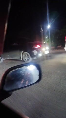 A point-of-view shot shows a vehicle traveling on a road at night. The reflection in the side mirror is prominent, revealing red and white lights, potentially from the dashboard or interior of the vehicle. Other vehicles pass by on the road. A white car is initially visible, followed by other cars and a green truck with multiple wheels and blue lights underneath. Streetlights illuminate the buildings and greenery along the roadside.