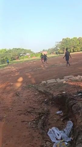In an outdoor setting under a clear sky, a group of young individuals is engaged in a casual soccer game on a dirt field. The playing surface is uneven, characterized by reddish-brown soil.  Around the perimeter of the field, there are trees and some residential structures visible in the distance. In the foreground, some exposed tree roots, along with scattered trash, suggest a somewhat unkempt or neglected area. The game is taking place in daylight, and the light appears to be moderate.