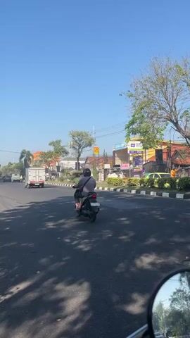 A street scene with clear blue skies and some clouds. Several motorcycles are traveling on the road. A white truck is also visible. Trees and buildings line the sides of the road. The video appears to be filmed from the perspective of someone riding on a motorcycle.