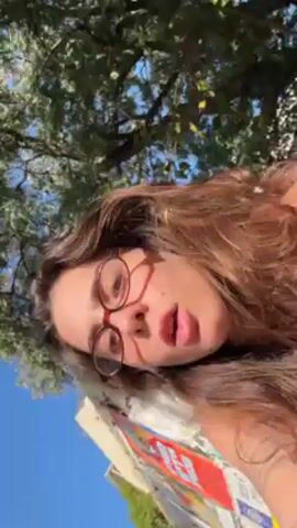 A woman with brown hair and glasses is outdoors on a sunny day. She is wearing a black tank top. The background includes trees, a building with white trim, a white umbrella, and other people seated at tables. The woman runs her hand through her hair, and then sticks her tongue out.