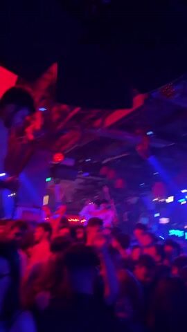 The video shows a crowded nightclub scene. The lighting is predominantly red and blue, creating a vibrant atmosphere. Many people are visible, mostly young adults, dancing and moving to the music. One individual is standing elevated above the crowd on some kind of structure, possibly a platform or bar, wearing a white t-shirt. The club has a complex ceiling with geometric shapes and lighting fixtures. Laser beams cut through the air, adding to the visual excitement. In the background, there's a sign that reads "TOP10". The camera appears to be hand-held.