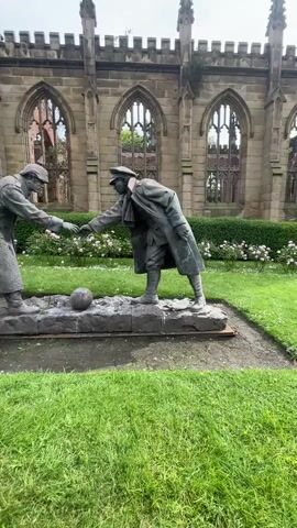 The video shows a statue depicting two soldiers reaching out to shake hands over a soccer ball, positioned in front of the ruins of a church. The soldiers are dressed in World War I era uniforms and greatcoats. Behind the statue is a partially ruined stone building with arched windows. There is a bed of flowers and some lawn in front of the building. The video zooms in closer on the statue. Other people can be seen moving in the background.