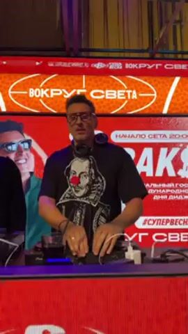 In a video capturing a DJ performance, a man wearing glasses and a black t-shirt adorned with a Shakespeare image and a red nose stands behind a DJ console, adjusting controls. His movements suggest he is actively mixing music. Beside him, to his left, stands another person wearing a dark coat. In the background is a large, red LED screen with the word "BOKPYT CBETA" displayed in white, the letters arranged in an oval shape around the words. Occasional blue stage lights illuminate the scene. A portion of a black laptop is visible to the DJ's right.