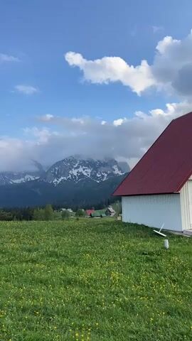 The video shows a scenic view of a mountainous landscape. The foreground features a lush green meadow dotted with yellow flowers, alongside a white building with a red roof. In the background, a mountain range is partially obscured by clouds. The sky is blue with scattered white clouds. The camera pans slightly, showing the full scope of the meadow and emphasizing the mountain range and the mix of coniferous and deciduous trees at the base of the mountains.  The scene evokes a sense of tranquility and natural beauty.